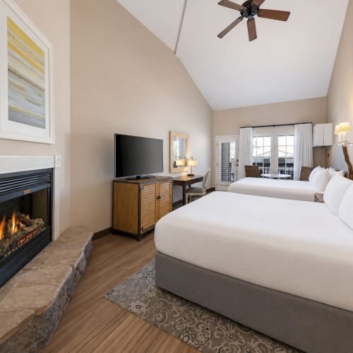 A bright modern hotel suite with two beds, a fireplace, a TV stand, and a ceiling fan.