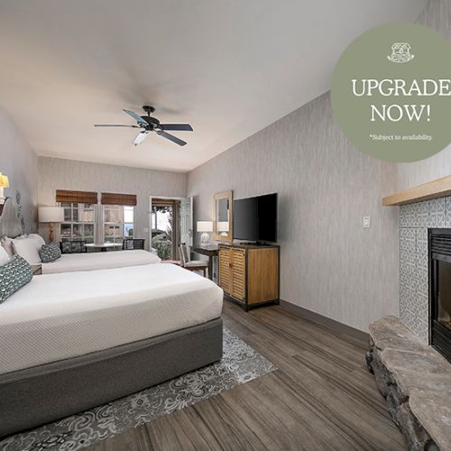 A modern hotel bedroom with a large bed, TV, ceiling fan, and a lit fireplace, with a &ldquo;UPGRADE NOW!&rdquo; promo banner.