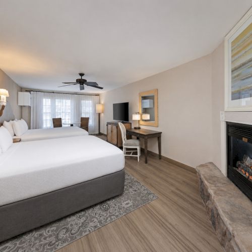 A modern hotel bedroom with two beds, ceiling fan, desk, and a lit gas fireplace in the corner.