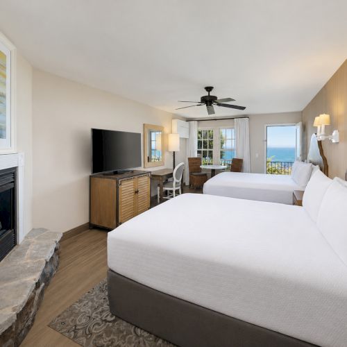 A bright hotel room with two beds, a ceiling fan, TV and console, a fireplace, and ocean-view windows.