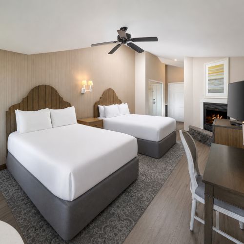 A tidy hotel-style bedroom with two queen beds, ceiling fan, fireplace, flat-screen TV, and a small desk area.