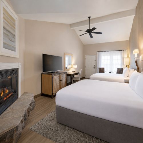Bright hotel room with two beds, fireplace, TV, lamps, ceiling fan, and windows with curtains.