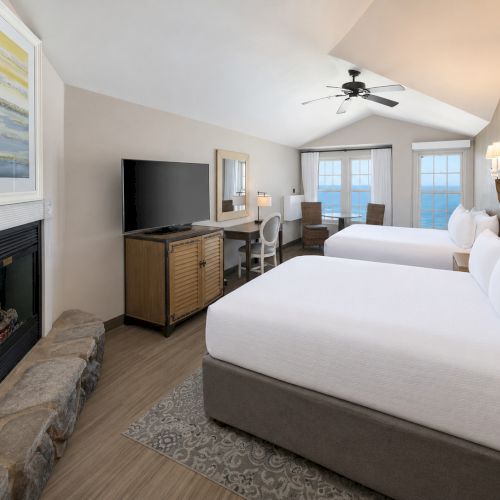 Bright hotel bedroom with two beds, a fireplace, TV on a cabinet, and ocean-view windows under a ceiling fan.