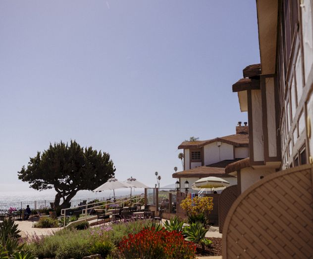 A coastal resort patio with umbrellas, flowers, trees, and a hotel overlooking the ocean under a cloudy sky.