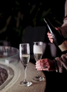 Two champagne flutes clink on a table as someone pours sparkling wine from a bottle during a toast celebration.