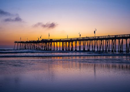 A long wooden pier with lamp posts stretches over the ocean at sunset, with the sky glowing in blue and orange clouds.