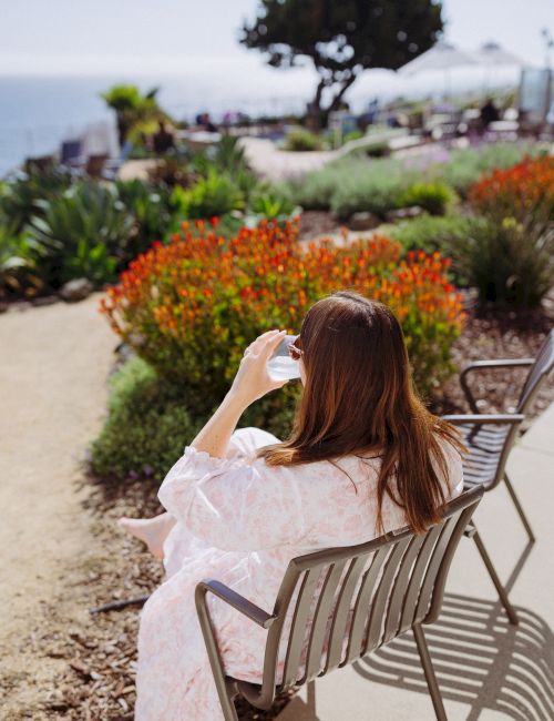 A woman relaxes in a patio chair, holding a drink as she gazes at a blooming garden of bright orange flowers.
