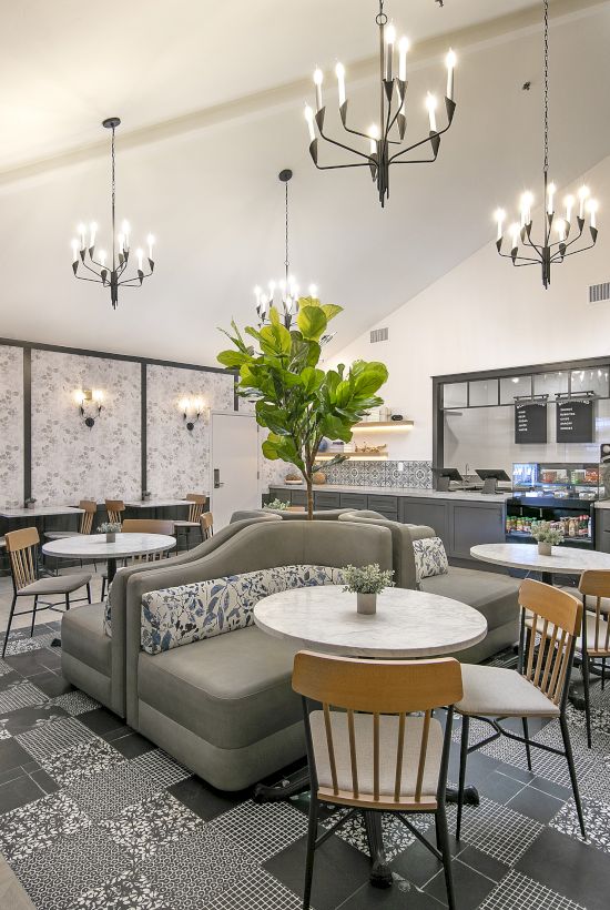 A modern caf&eacute; lounge with cushioned seating, round tables, hanging chandeliers, and a service bar in the background.