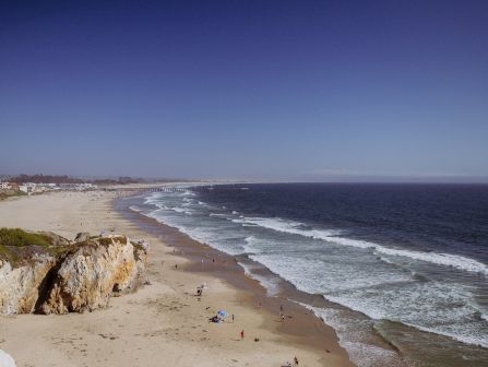 A wide sandy ocean beach with gentle waves, people relaxing, and a long pier stretching into the sea under a blue-gray sky.