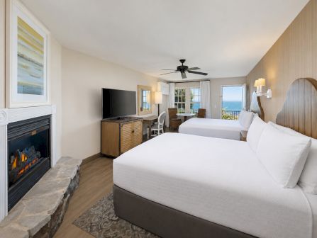A bright hotel bedroom with two beds, a ceiling fan, TV and fireplace, plus ocean-view windows.