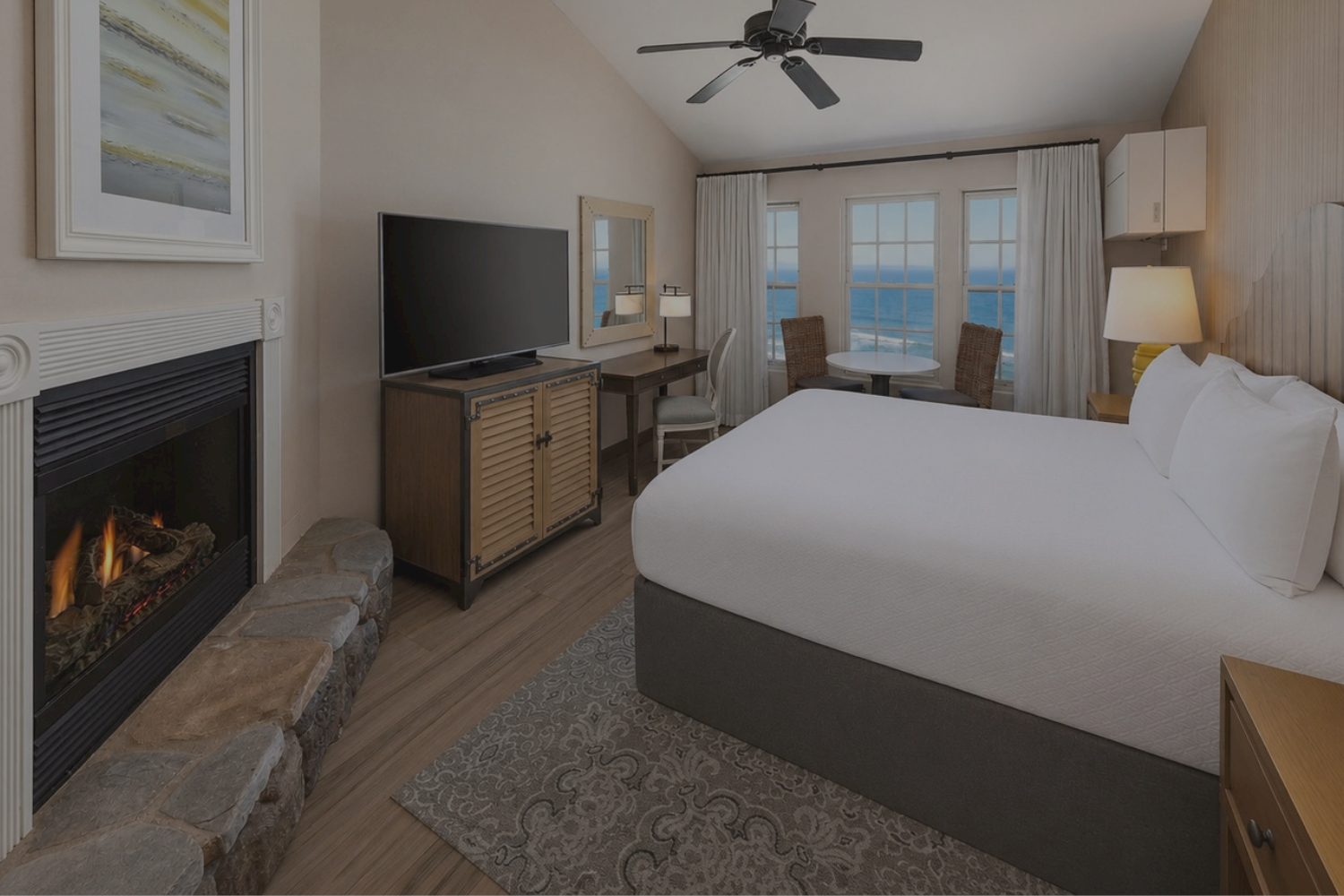 A modern hotel room with a king bed, TV, fireplace, ceiling fan, and ocean-view windows.