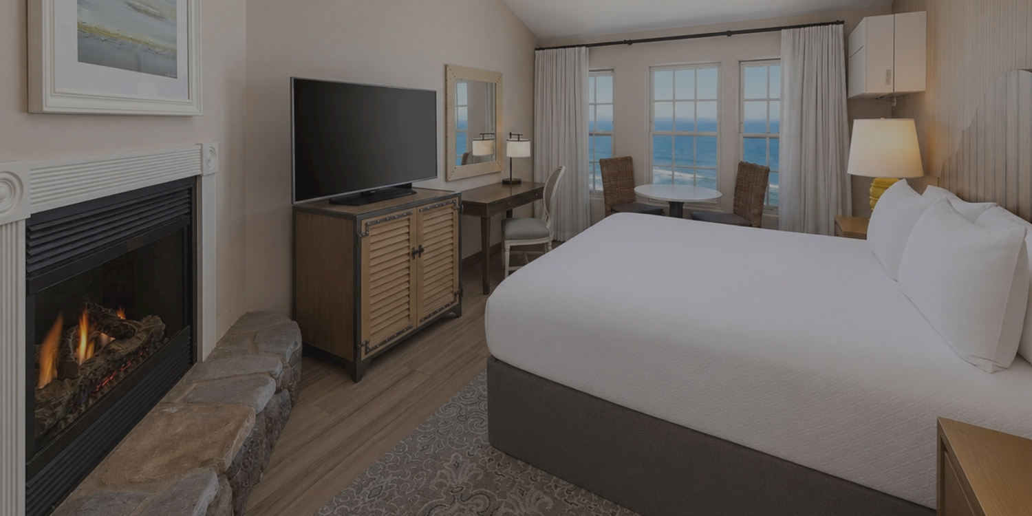 A modern hotel room with a king bed, TV, fireplace, ceiling fan, and ocean-view windows.