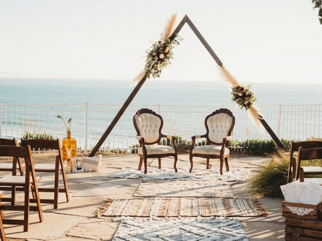 A beach wedding setup with two tufted chairs, floral arch, and candles on a sunlit terrace overlooking the ocean.