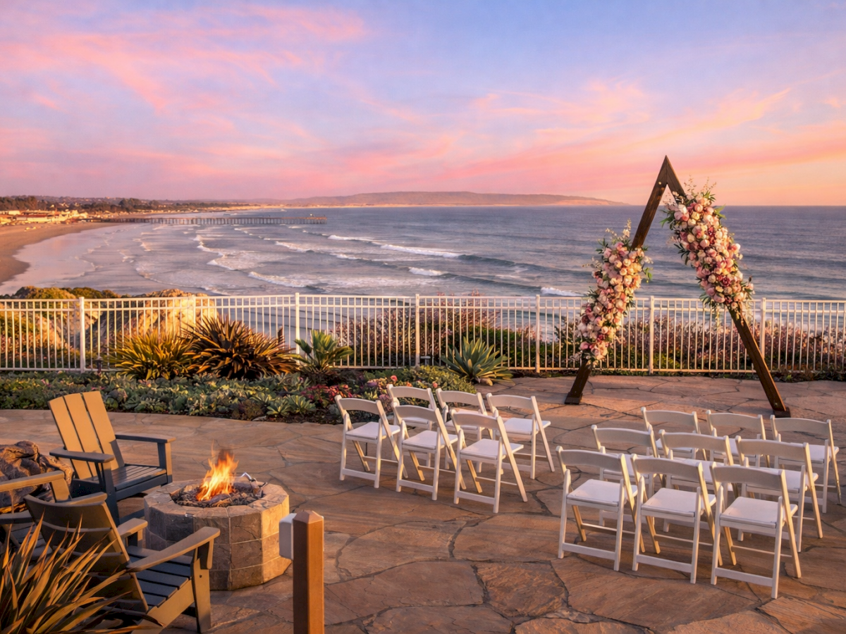 Beachside wedding patio at sunset with white chairs, a fire pit, and a floral arch by the ocean.