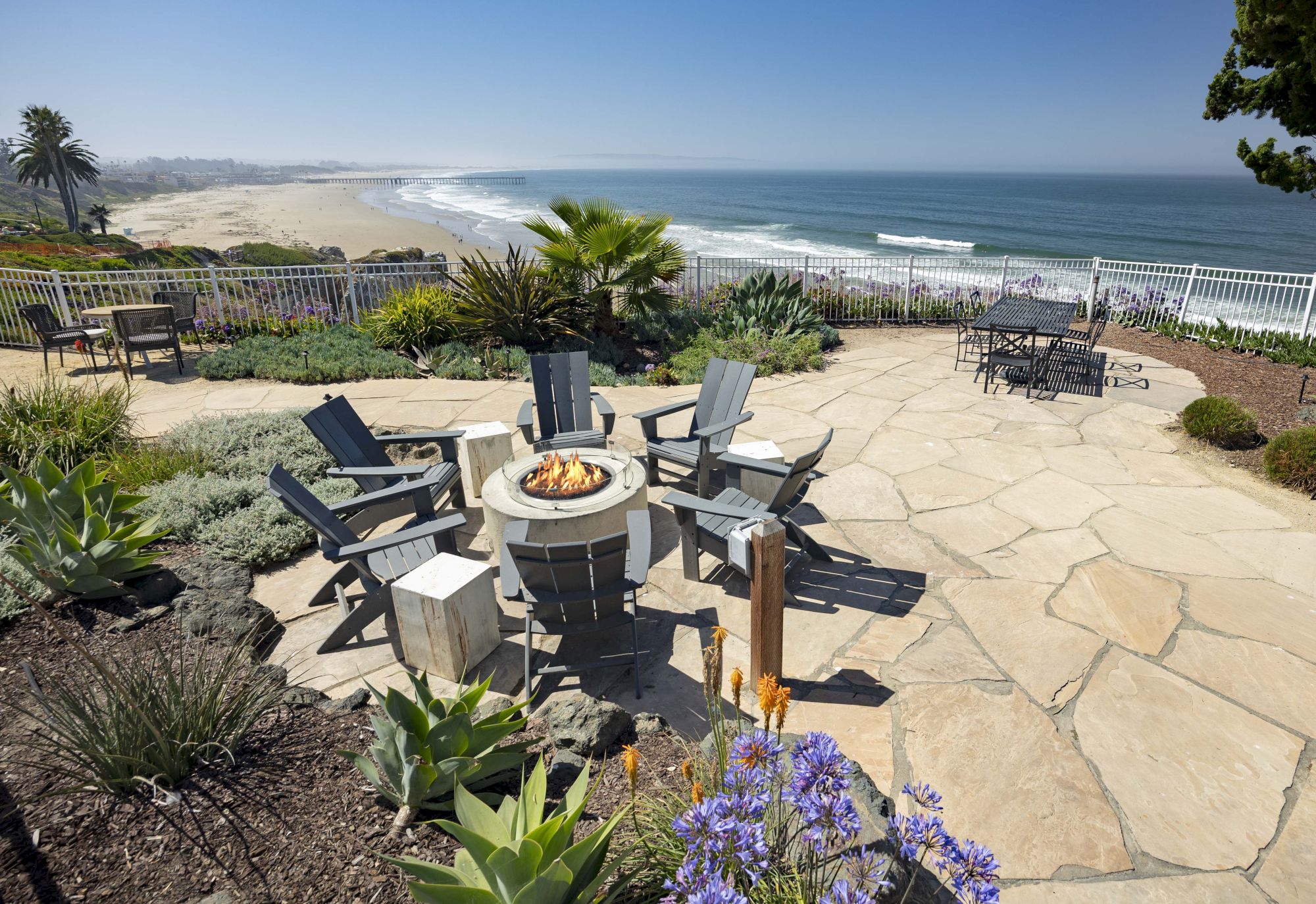 A seaside patio with a firepit, Adirondack chairs, palm plants, and a sweeping ocean view over a sandy beach.