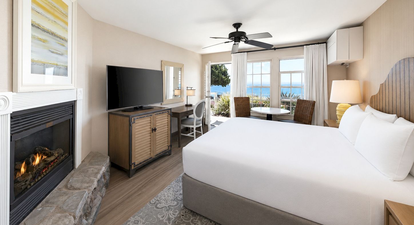 Modern hotel room with a fireplace, TV console, ceiling fan, and large windows overlooking the ocean.