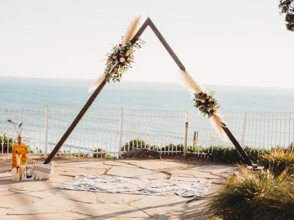 A beach wedding setup with a feather-and-flower arch, candles, and a scenic ocean view in the background.
