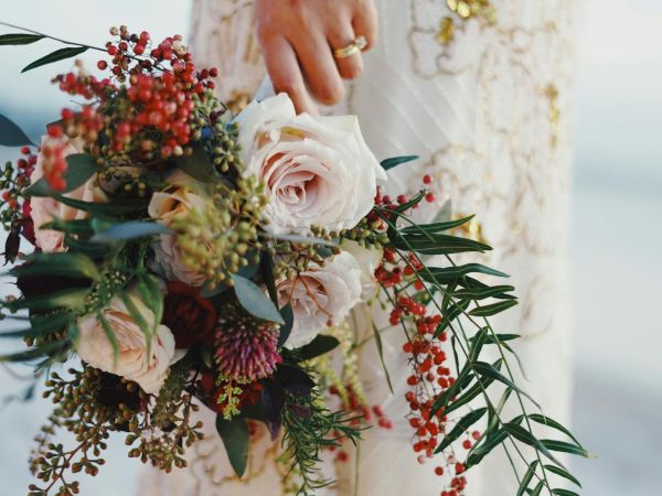 A bridal bouquet with white roses, red berries, and greenery, held by a bride in a lacy wedding dress by the water.