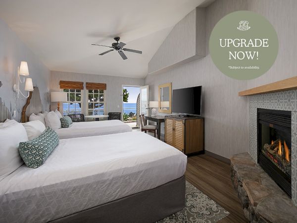 A modern hotel room with two beds, a TV and fireplace, plus a ceiling fan and a promo sign reading &ldquo;UPGRADE NOW!&rdquo;.